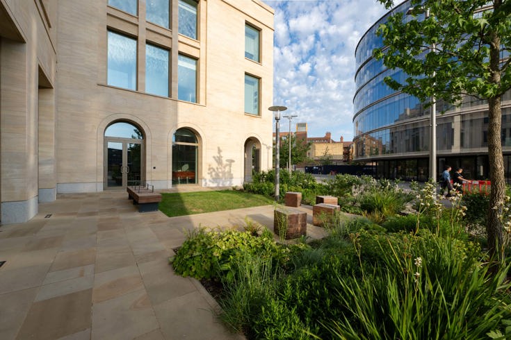 Landscaped courtyard with planting beds, seating and paved walkways between modern office buildings, featuring trees, shrubs and contemporary architecture.