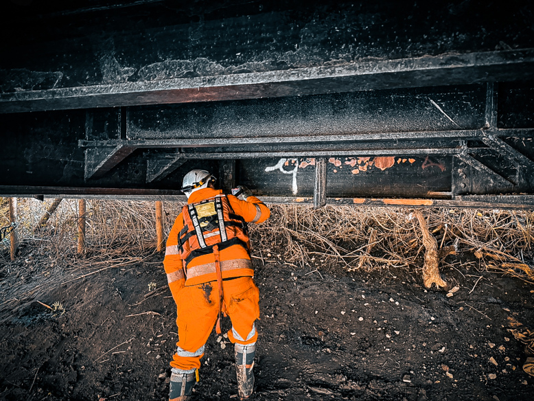 Worker in high-visibility orange inspecting the underside of a bridge structure.
