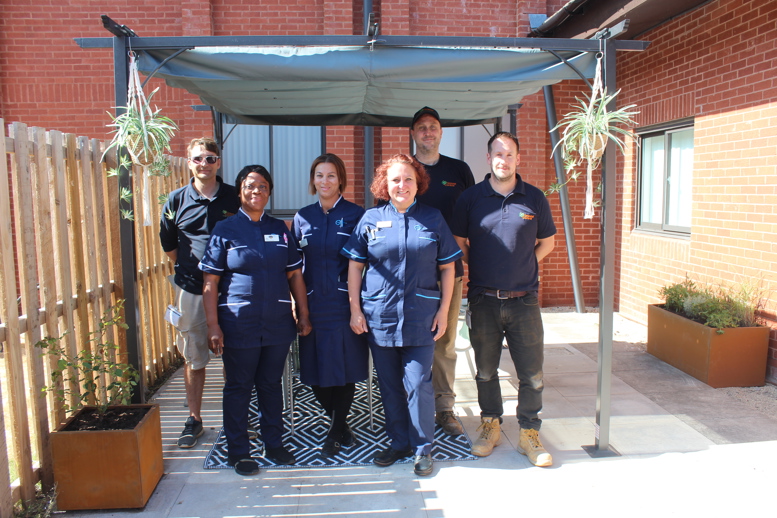 Care team and contractors standing beneath a shaded seating area in a landscaped outdoor courtyard.